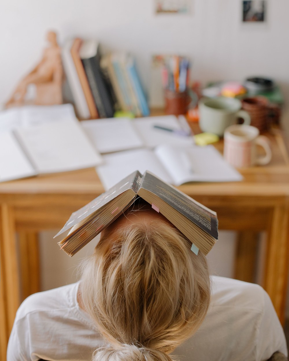 Studentin sitzt am Schreibtisch, den Kopf auf die Tischplatte gelegt, ein aufgeschlagenes Buch darauf — davor Notizen und Lehrbücher
